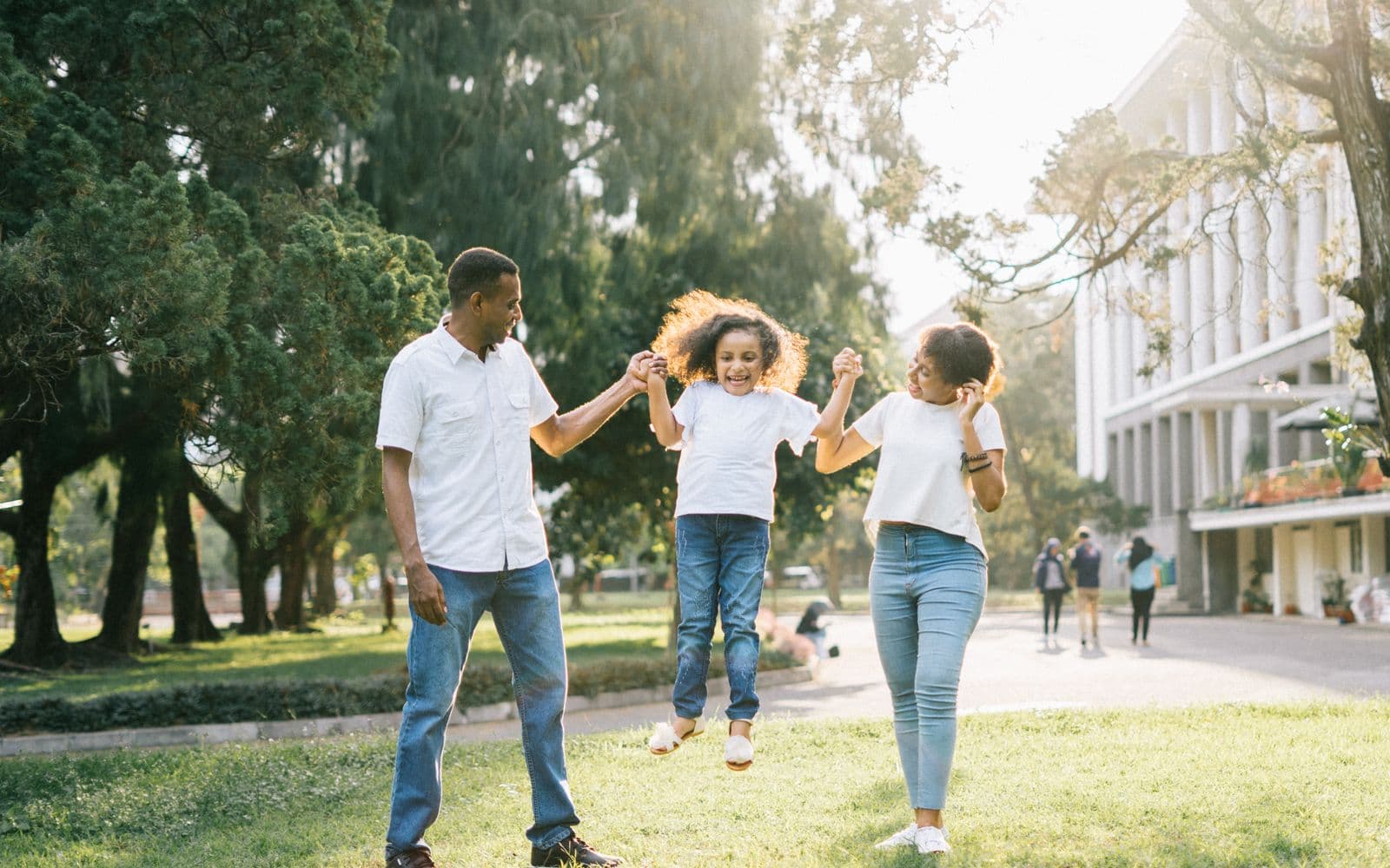 Joyful Black family swinging their young daughter outdoors on a sunny lawn